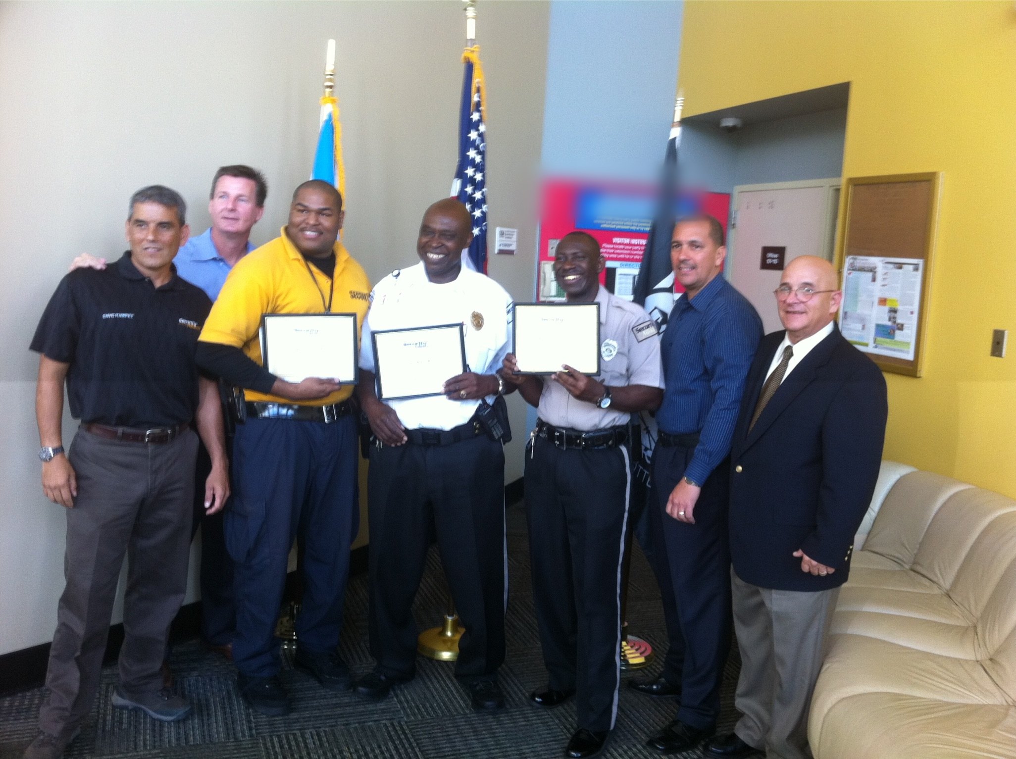 Three Security Alliance officers holding framed recognition certificates, standing with company leadership in an office lobby