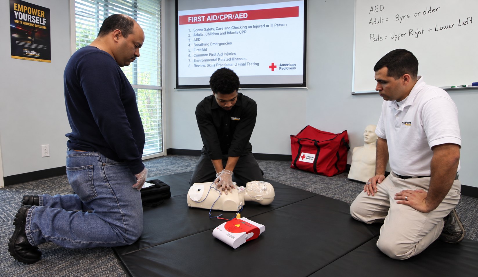 A trainee performing chest compressions on a CPR manikin during a first aid class, an AED and a red first aid bag beside the mat, with an instructor and a second trainee kneeling to observe