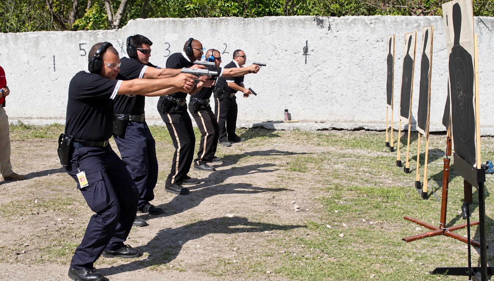 A line of Security Alliance officers in ear and eye protection firing pistols at paper targets on an outdoor range